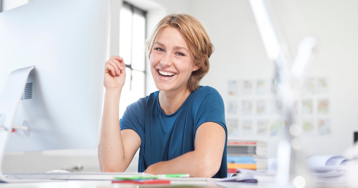 melhor curso para conseguir um emprego rápido - Retrato de mulher jovem feliz em sua mesa em um escritório criativo